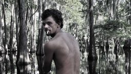 still, color image of shirtless Caucasian male with back facing camera and looking over his sholder with bayou in background take from documentary film 'The Other Side'